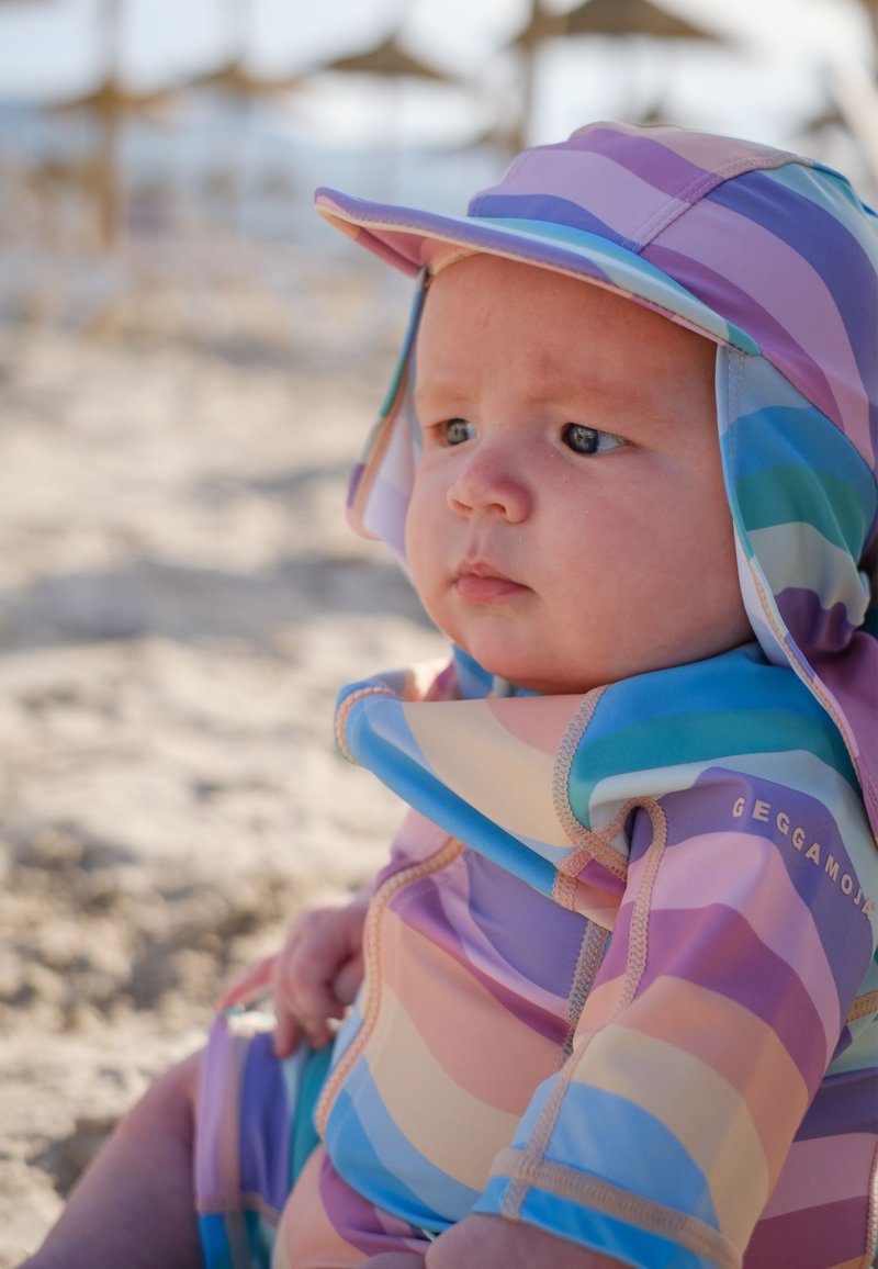 Värikäs uimapuku, jossa on leveät violettit, vaaleanpunaiset ja siniset raidat. Mukana on yhteensopiva aurinkohattu, jossa on lippa, suunniteltu auringolta suojaamiseen.