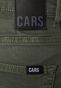Groene denimstof heeft een opvallend zwart label met "CARS" in het grijs, naast een kleiner zwart etiket met "CARS" in witte stiksel.