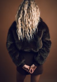 Brun fuskpälskort jacka med texturerade ärmar, bärs över en matchande ribbad brun kjol. Långt vågigt hår faller över ryggen.