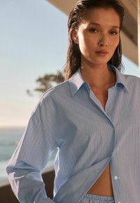 Camicia button-up azzurro chiaro con righe verticali, caratterizzata da un colletto classico e una vestibilità ampia. Il tessuto ha una texture morbida, che amplifica il suo aspetto informale.