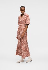 Mujer que lleva un vestido largo con cinturón, estampado floral y color óxido, de manga corta, de pie y mirando hacia abajo, combinado con zapatos planos beige claro.