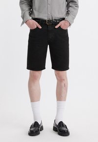 Shorts de mezclilla negra con corte recto, combinados con calcetines blancos hasta la altura del tobillo y mocasines negros. El conjunto incluye una camisa de botones gris claro y un cinturón trenzado.