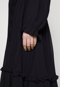 Abito nero plissettato con maniche lunghe e polsini arricciati, caratterizzato da un design fluido e tessuto morbido e leggero. Mano posizionata sull'abito.