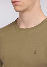T-shirt z bawełny w oliwkowym kolorze, z okrągłym dekoltem. Posiada subtelnie wyszywane logo po lewej stronie klatki piersiowej. Gładka tekstura, dopasowany krój.