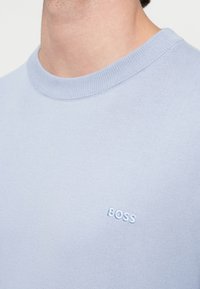 Κοντινό πλάνο ενός ατόμου που φορά ένα ανοιχτό μπλε πουκάμισο με στρογγυλή λαιμόκοψη και κεντημένη τη λέξη "BOSS" στο στήθος.