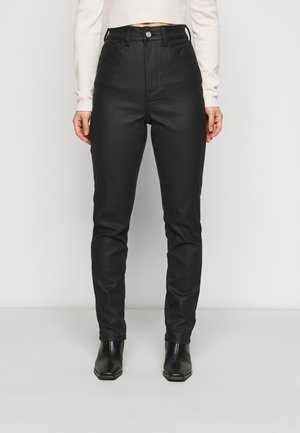 Person iført sorte læderbukser med høj talje og sorte ankelstøvler, kombineret med en cropped hvid sweater med lange ærmer, stående på en neutral baggrund.