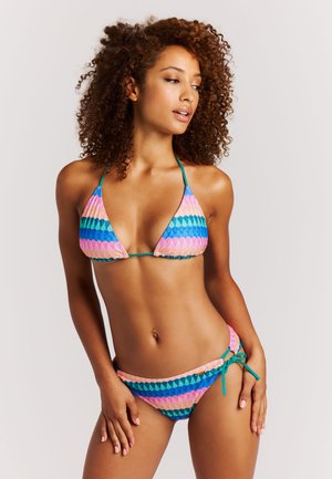 Vrouw met krullend haar die een roze, blauwe en groene gestreepte bikini draagt met een haltertop en zijstriksluiting aan de onderkant, poserend tegen een effen lichte achtergrond.