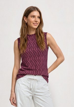 Femme aux longs cheveux bruns portant un haut violet sans manches à motifs et un pantalon blanc à cordon, debout devant un fond clair uni.
