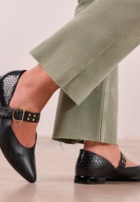 Zapatos negros de puntera afilada con un acabado texturizado de reptil, una correa con hebilla y tachuelas doradas, y un tacón brillante, combinados con pantalones verdes cortos.