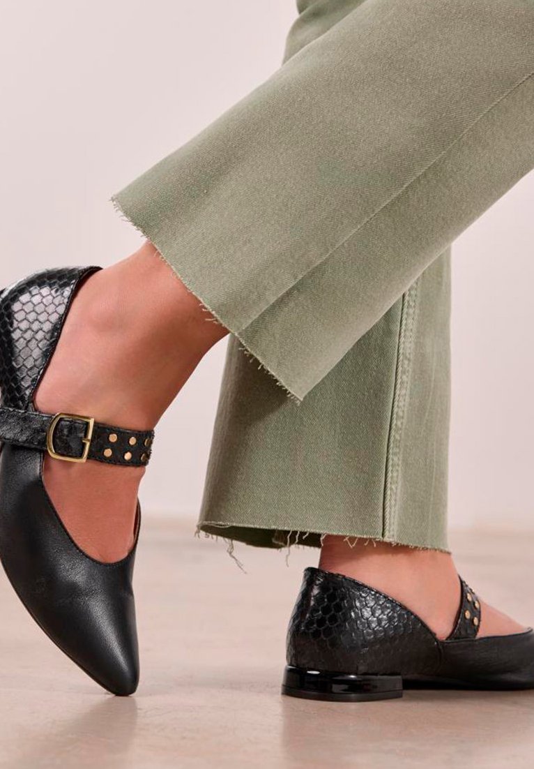 Zapatos negros de puntera afilada con un acabado texturizado de reptil, una correa con hebilla y tachuelas doradas, y un tacón brillante, combinados con pantalones verdes cortos.