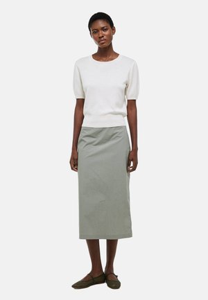 Femme portant un pull à manches courtes beige clair, une jupe midi grise et des chaussures plates vert foncé, debout devant un fond uni.