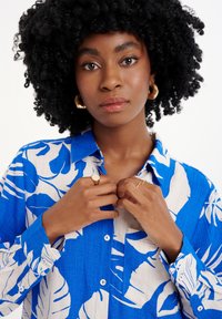 Camicia azzurra e beige con motivo floreale, patta con bottoni e maniche lunghe, realizzata in tessuto testurizzato. Orecchini a cerchio dorati e anelli.