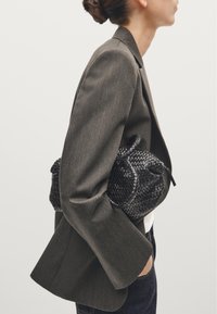 Zwarte geweven handtas met een ronde vorm, vastgehouden tegen een aansluitende grijze blazer. De blazer heeft een textuur stof met een subtiele verticale streep.