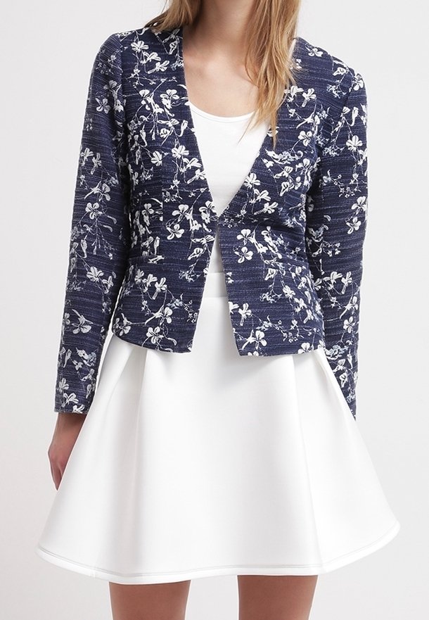 Femme portant un blazer cintré bleu marine à motifs floraux sur une robe blanche sans manches avec une jupe évasée.