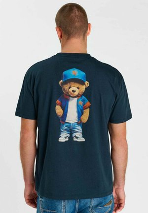 Uomo che indossa una t-shirt scura con la grafica di un orsetto vestito con una giacca blu, cappellino, camicia bianca, jeans e sneakers sul retro.