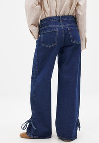 Široke temno modre denim hlače z visokim pasom, zadnjimi žepi in nastavljivimi vezalkami na robu. Gladka tekstura z diskretnimi šivi.