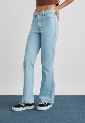 Persoon draagt lichtblauwe rechte jeans en zwarte sneakers met witte zolen, staand op een blauw tapijt tegen een effen muur.