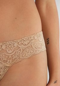 Lace bikini slip in een nude kleur met een bloemenpatroon op de tailleband, voorzien van een zachte textuur en elastische zijkanten voor comfort.