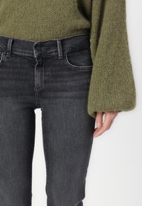 Jean gris foncé avec une coupe slim, featuring des poches avant et une fermeture à bouton, associé à un pull en maille texturée de couleur olive.