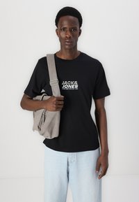 Camiseta de algodón negra con logo impreso en blanco, combinada con jeans de mezclilla azul claro y un bolso gris de hombro con múltiples bolsillos.