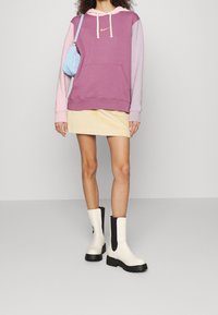 Lila och rosa färgblockerad hoodie med framficka, kombinerad med en beige kordfläta mini kjol och vitt-svarta ankelstövlar.
