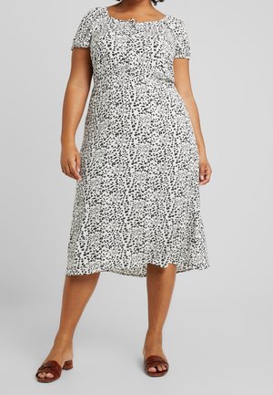 Kobieta w białej sukience midi z czarnym kwiatowym wzorem i krótkimi rękawami, w zestawieniu z brązowymi sandałami na niskim, szerokim obcasie z odkrytymi palcami.