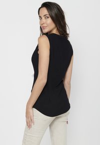 Schwarzes ärmelloses Top mit tailliertem Design, rundem Saum und weicher Textur. Getragen mit hellen Jeans, die minimalistische Details aufweisen.