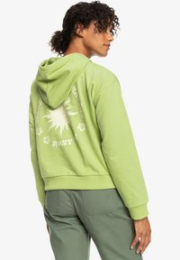 Hoodie cropped verde chiaro con coulisse, caratterizzato da una grafica del sole e accenti floreali sul retro. Tessuto morbido e testurizzato con polsini a coste.
