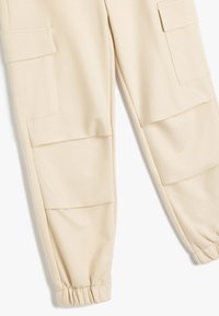 Beige cargo bukser med en afslappet pasform, elastiske ankelmanchetter og to store sidelommer med klaplukninger. Glat stoftekstur.