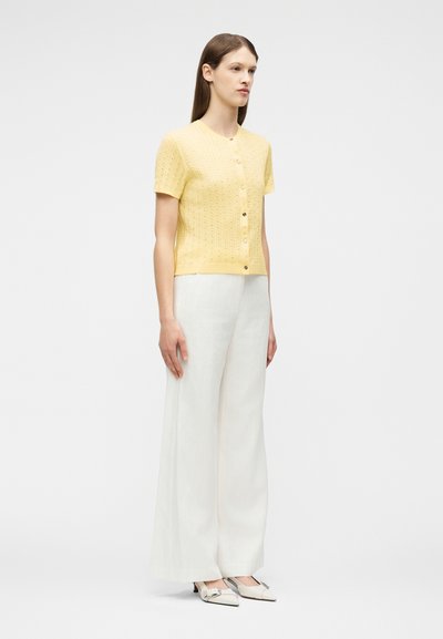 Γυναίκα που στέκεται, φορώντας ένα κίτρινο πουλόβερ με κουμπιά και κοντά μανίκια, λευκό παντελόνι με φαρδιά μπατζάκια και λευκά μυτερά παπούτσια με φιόγκους.