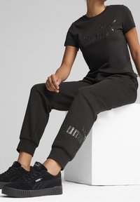 Schwarzes Baumwoll-T-Shirt und passende Sweatpants, beide mit einem strukturierten schwarzen Grafikdesign. Kombiniert mit schwarzen Wildledersneakern, die auf einem weißen Würfel stehen.