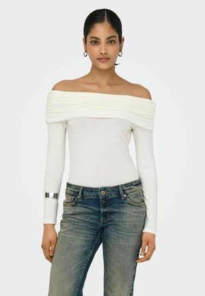 Hvid off-shoulder top i struktureret stof, lange ærmer, parret med blå denimjeans. Sølvarmbånd accentuerer det ene håndled.