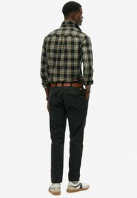 Homme portant une chemise à carreaux verte et jaune, un pantalon noir avec une ceinture marron et des baskets blanches, debout les mains dans les poches, tourné le dos.