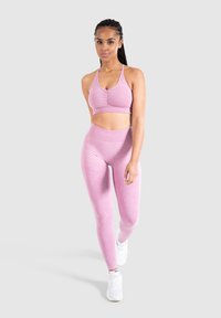 Top sportivo rosa chiaro con dettagli arricciati, abbinato a leggings rosa a vita alta. Entrambi hanno una texture liscia. Sneakers bianche completano il look.