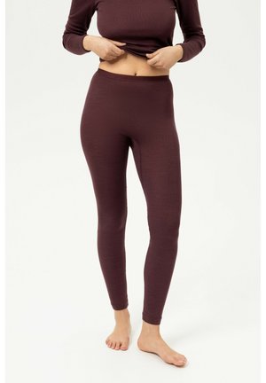 Vrouw draagt een donkerbruine top met lange mouwen en bijpassende legging, tilt haar shirt iets op om haar blote buik te tonen, staat op blote voeten.