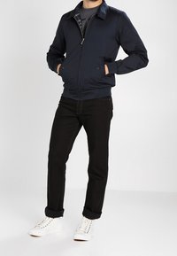 Marinblå bomberjacka med dragkedja framtill, ribbade muddar och två sidofickor, kombinerad med svarta jeans och vita sneakers. Enkel design, utan mönster.