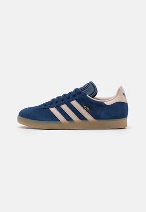 Niebieski zamszowy sneaker Adidas Gazelle z beżowymi paskami, gumową podeszwą i logo Adidas na pięcie, przedstawiony z boku na białym tle.