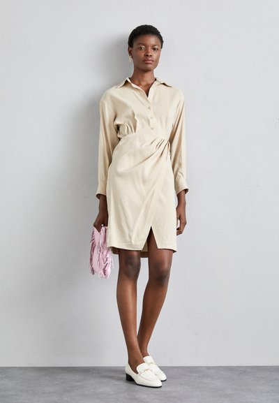 Vestido beige de botones con mangas largas, cintura ceñida y una abertura frontal. Combinado con mocasines blancos y un bolso de mano con flecos en rosa.