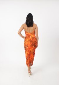 Bagfra af en lang, tætsiddende kjole i strålende orange med blomsterdesign; har tynde stropper og en sideslids for nem bevægelse.