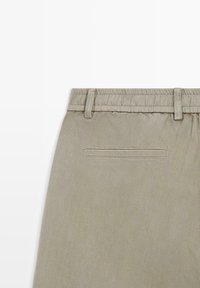 Beżowe spodnie z elastycznym pasem, szlufkami na pasek oraz pojedynczą wsuwaną kieszenią z tyłu na białym tle.