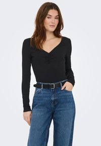 Sort langærmet top med V-halsudskæring og samlet detalje. Kombineret med højtsiddende blå denimjeans og sort bælte.