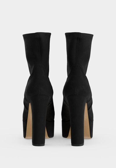 Paire de bottines noires à talons hauts avec talons blocs, vues de dos sur un fond clair uni.