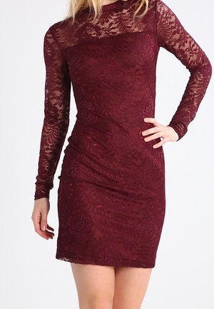 Frau in einem eng anliegenden, langärmeligen, burgunderroten Spitzenkleid mit Blumenmuster, die mit einer Hand in der Hüfte vor einem weißen Hintergrund steht.