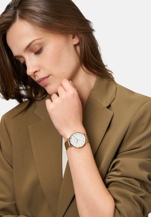 Montre en maille dorée avec un cadran texturé argenté, présentant des marqueurs d'heure minimalistes. Portée au poignet, associée à un blazer marron.