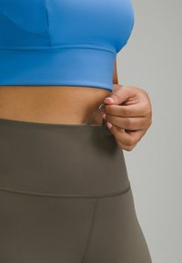 Blauwe braf top, gemaakt van gladde stof, gecombineerd met olijfgroene high-waisted leggings. Een hand past een kleine band aan de tailleband.