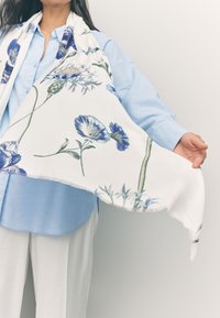 Foulard floral avec un fond blanc, présentant des fleurs bleues et des tiges vertes, en tissu doux avec des bords effilochés, tenu par une personne.