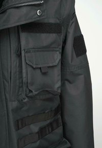 Fekete taktikai dzseki sima textúrával, több zsebbel, tépőzáras felvarrókkal és vízszintes hevederszíjakkal a rögzítéshez.