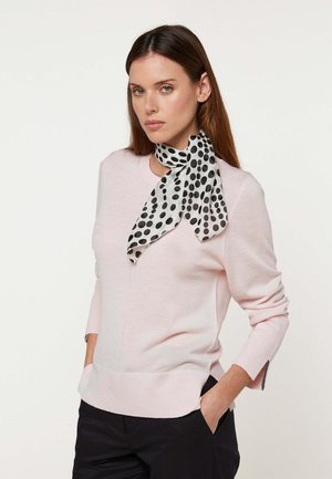 Frau mit glattem braunem Haar trägt ein hellrosa langärmeliges Oberteil, schwarze Hose und einen weißen Schal mit großen schwarzen Punkten, der um den Hals gebunden ist.