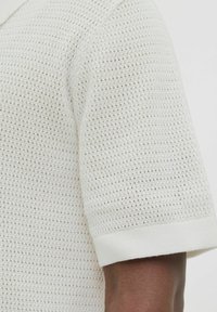 Bijela textured polo majica s uzorkom mreže, rebrastim manžetama i glatkom završnom obradom, prikazuje rame i dio rukava.