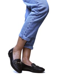 Osoba w podwiniętych niebieskich dżinsach i czarnych plecionych mokasynach z srebrnymi zapięciami, pozująca na białym tle.
